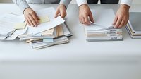 Male professional in business attire organizing and sorting stacks of documents and paperwork on white desk, managing files and administrative tasks in office environment.