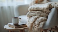 Cozy home atmosphere with knitted beige sweater on armchair, coffee cup and books on round table. Soft morning light through curtains creates peaceful mood.