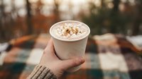 Hot chocolate with marshmallows and cinnamon in paper cup held by hand wearing knitted sweater on cozy autumn background for seasonal drinks.