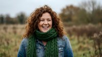Happy middle-aged Caucasian woman with curly red hair wearing green scarf and denim jacket smiling outdoors in autumn countryside field landscape.