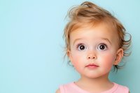 Adorable toddler with big brown eyes and wispy blonde hair wearing pink against light blue background, expressing wonder and curiosity in studio portrait setting.