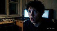 Young man with curly dark hair and fair complexion, wearing glasses, sitting in dimly lit room with computer monitors and vintage equipment in background, looking directly at camera with serious expression.