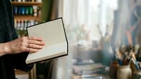 Person holding open blank notebook with cityscape view through window, suitable for education, business planning, creative writing, and lifestyle concepts.