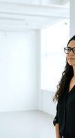 Young woman with long dark hair and glasses wearing black blazer standing in bright, minimalist white office space for corporate, business, and professional portraits.