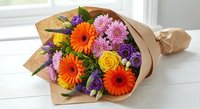Vibrant mixed flower bouquet with orange gerberas, purple roses, yellow roses and pink chrysanthemums wrapped in brown kraft paper on white surface.