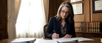 Professional woman with long dark hair and glasses, wearing dark blazer, writing at wooden desk in law office with bookshelves, documents, and window with curtains.