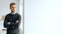 Young Caucasian male professional wearing dark sweater standing with crossed arms in modern office space with white walls and natural lighting.