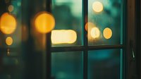 Atmospheric evening window view with glowing bokeh lights creating dreamy mood. Raindrops on glass pane reflect warm golden circles against dark teal background.
