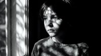 Young woman with dark hair looking pensively through window with dramatic shadows and light patterns creating moody black and white portrait atmosphere.