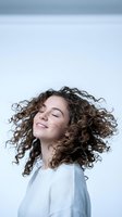 Young woman with curly brown hair smiling joyfully with eyes closed against white background. Natural beauty and happiness concept for wellness campaigns.