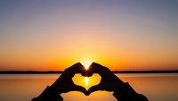 Hands forming heart shape silhouette against stunning sunset over calm water creating romantic peaceful moment for love wellness meditation concepts.