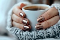 Female hands with sparkly rose gold glitter manicure holding white coffee cup, wrapped in cozy gray knitted sweater, creating warm winter morning atmosphere.