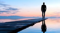 Silhouette of lone man standing on wooden pier over calm lake at sunset, with mountains in background and reflection in water creating symmetrical scene.