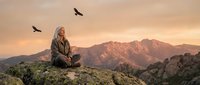 Elderly man with long gray hair meditating on mountain peak at sunset with soaring birds overhead in peaceful wilderness landscape for mindfulness concepts.