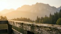 Wooden fence railing in sharp focus with blurred mountain landscape and evergreen forest at sunrise or sunset, suitable for nature and outdoor design projects.