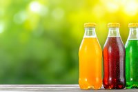 Colorful glass bottles with orange, red and green carbonated beverages on wooden surface against blurred natural bokeh background. Fresh summer drinks.