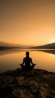 Woman meditating in lotus position on rock by calm lake at golden sunset, mountains reflected in water, mindfulness and wellness concept.