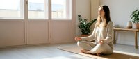 Young woman of East Asian descent in casual beige clothing practicing meditation or yoga on a mat in a bright, minimalist room with natural light from large windows and potted plants.