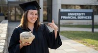 Young Caucasian female graduate in cap and gown holding education fund piggy bank near university financial services building, celebrating debt-free education.