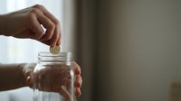 Hand dropping coin into glass jar for savings and financial planning goals with soft natural lighting and blurred background for copy space.