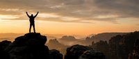 Silhouette of successful man with raised arms celebrating victory on mountain peak at golden sunrise with dramatic cloudy sky and rocky landscape below.