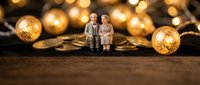 Miniature elderly couple figurines sitting on golden coins with warm bokeh lights background representing retirement savings and financial planning.
