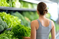 Young Caucasian woman in gray tank top choosing fresh green lettuce and vegetables at grocery store produce section, view from behind, healthy lifestyle concept.