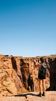 Female hiker with backpack standing at rocky canyon cliff edge under clear blue sky, exploring arid desert landscape and geological formations during outdoor adventure.