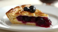 Fresh baked cherry and blueberry pie slice with flaky golden pastry crust and creamy custard filling, garnished with whole berries on white plate.