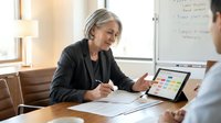 Mature woman with gray hair and Caucasian ethnicity, wearing black blazer, conducts business meeting or coaching session with male colleague in modern office, reviewing documents and tablet for corporate training, consulting, or mentoring purposes.