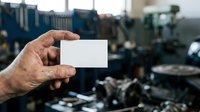 Worker holding blank white business card at industrial construction site with machinery and equipment in background for branding and corporate identity mockup.