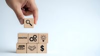 Hand placing wooden cube with magnifying glass icon completing business strategy puzzle with analysis, budget, handshake and dollar symbols on white background.