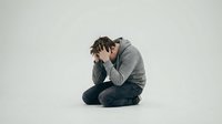 Young man in gray hoodie sitting in despair with head in hands showing mental health struggles and emotional distress on neutral background.