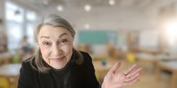Senior female teacher with gray hair smiling and waving in classroom with desks and chalkboard in background, creating welcoming educational atmosphere.