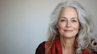 Elegant mature woman with long silver hair and natural makeup wearing burgundy sweater smiles warmly against neutral background, radiating confidence and grace.