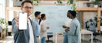 Professional businessman with glasses displaying blank smartphone screen in modern office meeting room with diverse team collaborating on whiteboard project.