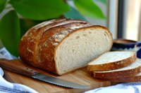 Freshly baked artisan sourdough bread loaf sliced on wooden cutting board, showing soft white crumb texture and crispy golden brown crust with flour dusting.