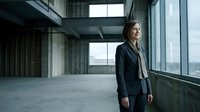 Middle-aged woman with long brown hair, Caucasian ethnicity, wearing dark business blazer and light shirt, standing in modern empty office space with concrete floors and large windows overlooking cityscape. Suitable for corporate, business leadership, professional development, and workplace concepts.