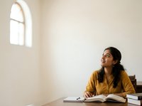 Young woman with long dark hair and South Asian ethnicity, wearing a mustard-yellow shirt, studying at a wooden desk with an open book in a bright, minimalist room with arched window. Educational and learning concept.