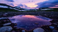 Mountain lake reflects dramatic purple and pink sunset sky, with snow patches on distant peaks and rocky shore creating serene alpine wilderness landscape.