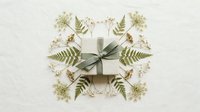 Elegant gift box with green ribbon surrounded by delicate fern leaves and small white flowers on neutral background for special occasions.