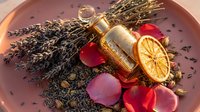 Luxury aromatherapy setup with lavender bundle, golden essential oil bottle, dried orange slice and rose petals in ceramic bowl for spa relaxation.