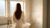 Young woman in white bathrobe standing by luxury bathtub in bright bathroom with natural sunlight streaming through window creating peaceful morning atmosphere.
