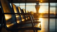 Empty airport terminal seating area at sunset with warm golden light streaming through windows, casting long shadows across modern black chairs in waiting area.