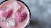 Pink strawberry milkshake with heart shaped candy decorations and ice cubes in glass, romantic beverage close up on blurred background, selective focus.