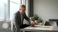 Middle-aged Caucasian male professional in business casual attire working at modern office desk with laptop and potted plant, natural window lighting.