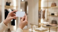 Hands holding blank white card in luxury retail store with fashion displays and shelves in background.