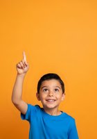 Young boy with dark hair and light skin tone wearing bright blue t-shirt, smiling with raised index finger pointing upward against vibrant orange background for educational and advertising campaigns.