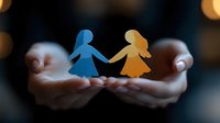 Paper cutout silhouettes of two female figures holding hands in cupped palms against dark background with bokeh lights, symbolizing friendship, support and unity.