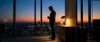 Professional businessman silhouette working late in modern office with city skyline view during sunset. Corporate executive at desk with lamp in skyscraper.